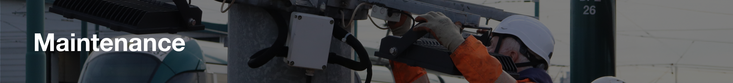 On the left is an older man with a neon orange safety jacket on with reflective stripes. On his head is a white hard hat with black straps and he had on navy trousers with a blue harness around his thighs. He is doing works on a large silver mast. On the right of the image is two workers being yielded up to do maintenance work on the lighting structure 
