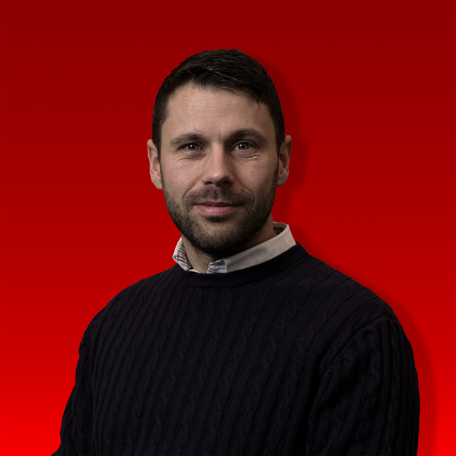 Dan Haystead sat down with navy knit jumper on and shirt sticking out of collar. Dark brown hair and small amount of facial hair 